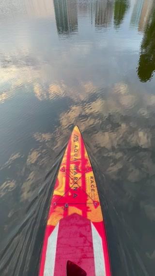 A red and orange paddleboard floats on a calm body of water reflecting clouds